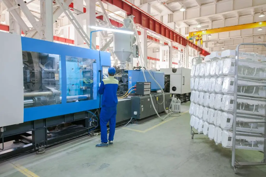 Worker operating a blue injection molding press beside a pallet of finished parts